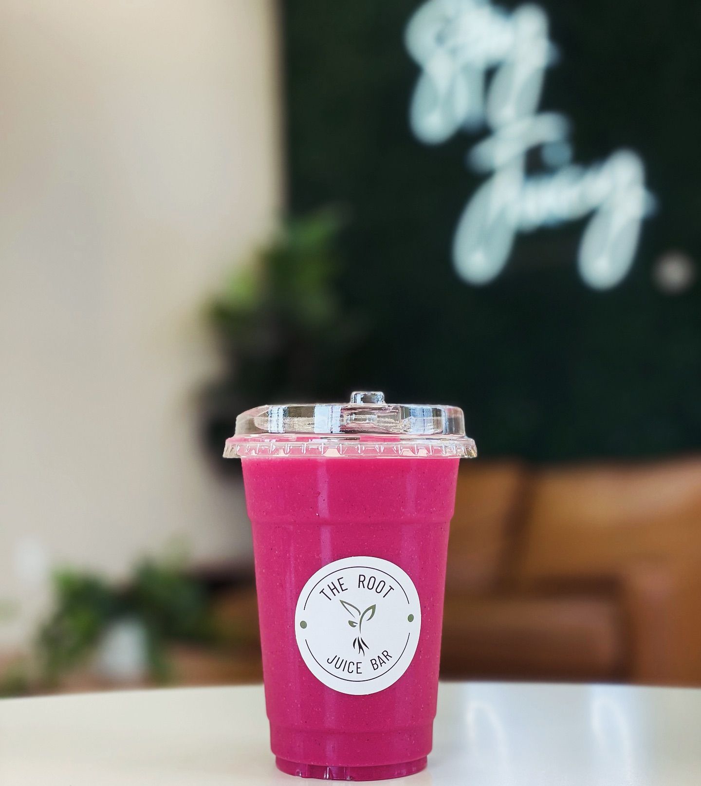 A bright pink smoothie in a clear plastic cup with a circular logo, sitting on a table in front of a blurred shop sign.