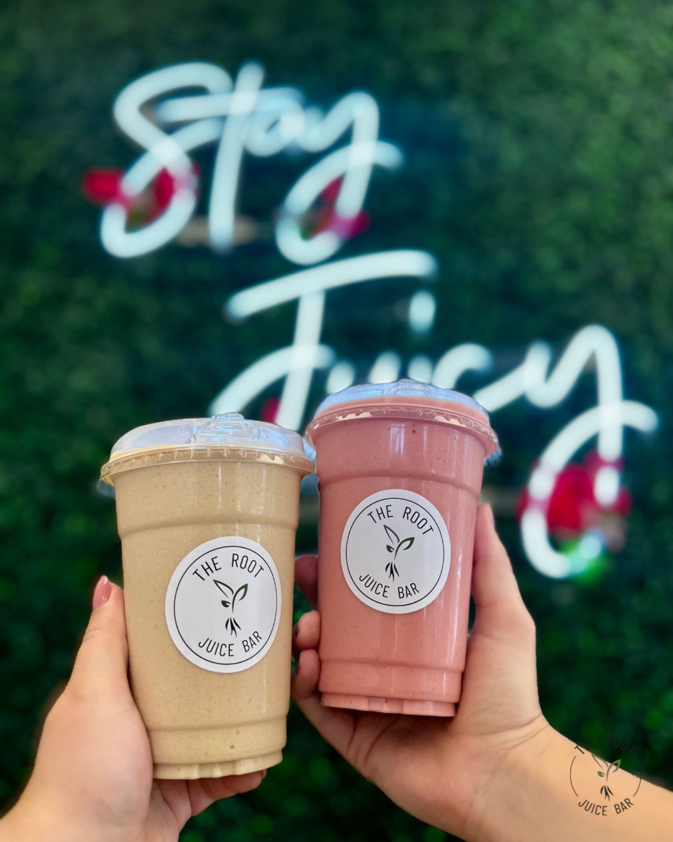 Two hands hold up beige and pink smoothies in front of a neon 
