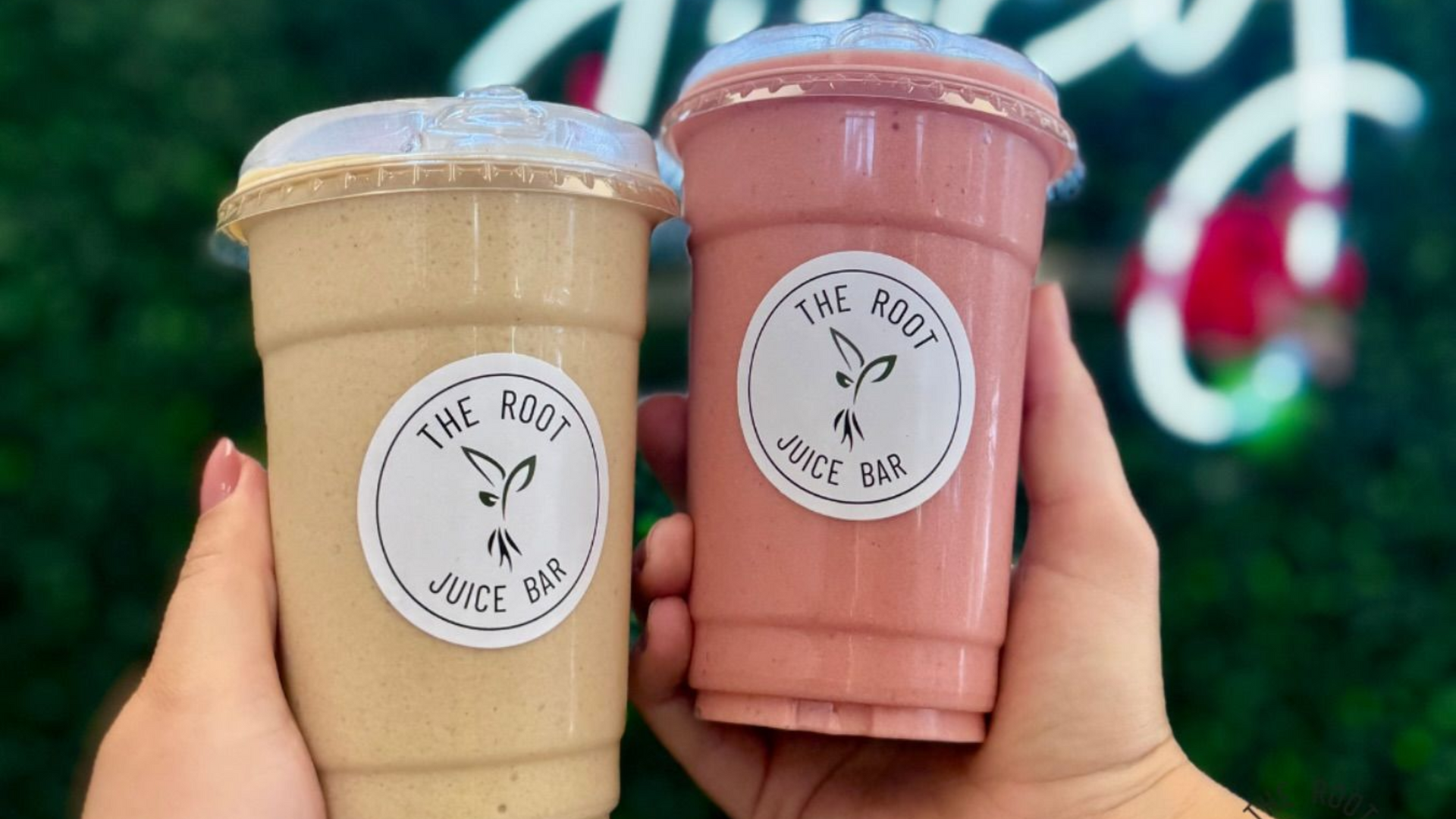 Two hands hold a tan smoothie and a pink smoothie, both featuring The Root Juice Bar labels, against a green background.