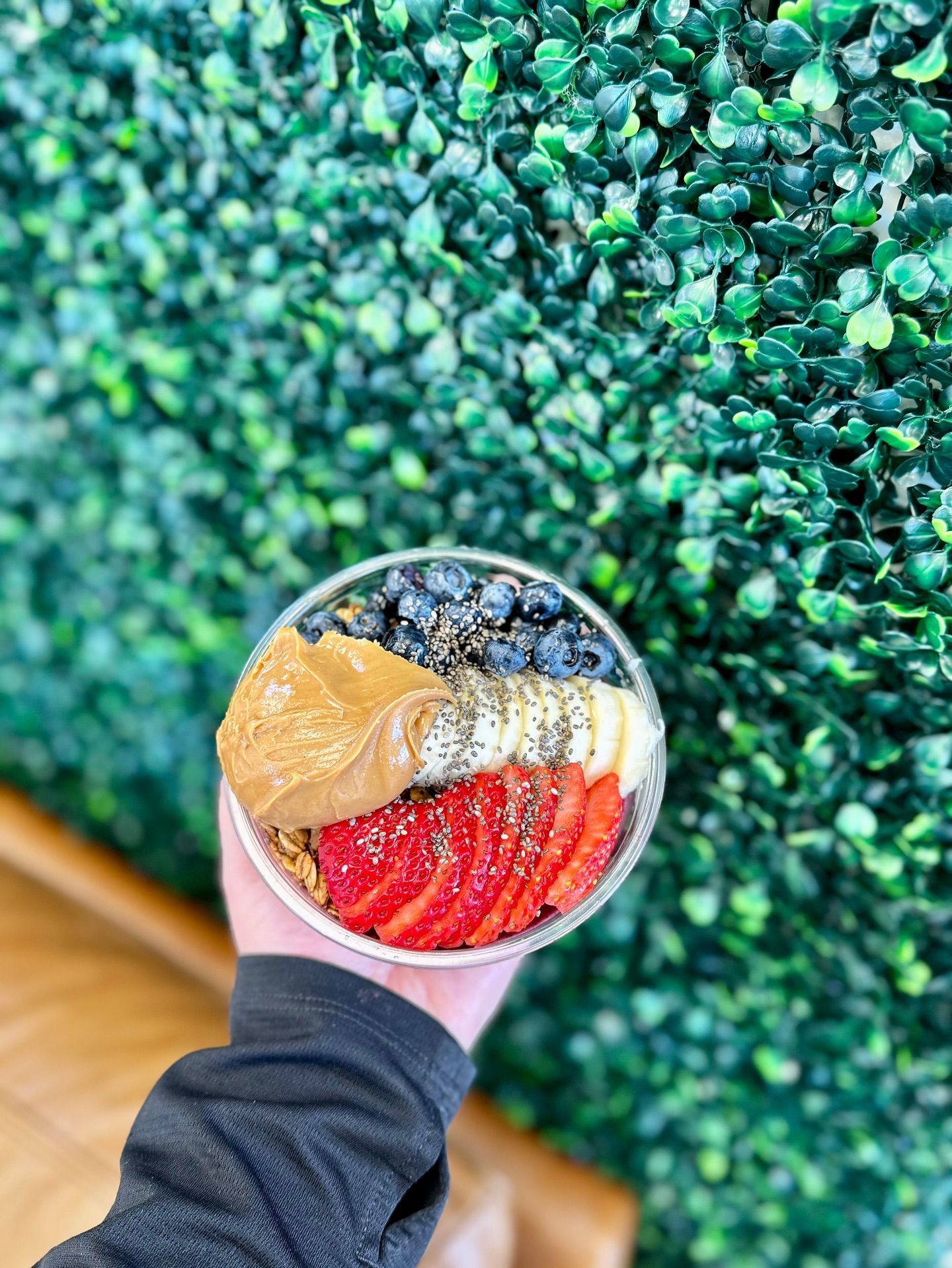 A hand holds a smoothie bowl topped with sliced strawberries, banana, peanut butter, blueberries, and seeds against a wall.