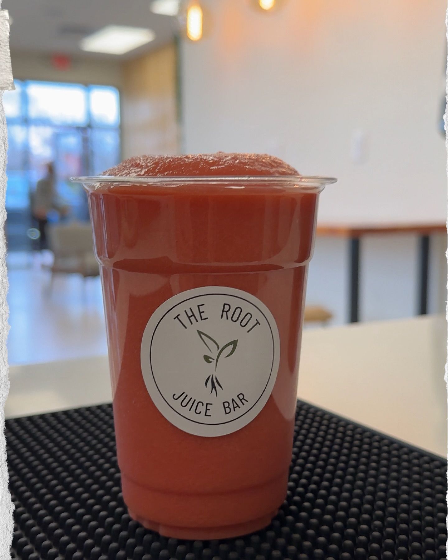 A plastic cup of reddish-pink smoothie from The Root Juice Bar, sitting on a black bar mat in a bright, modern shop.