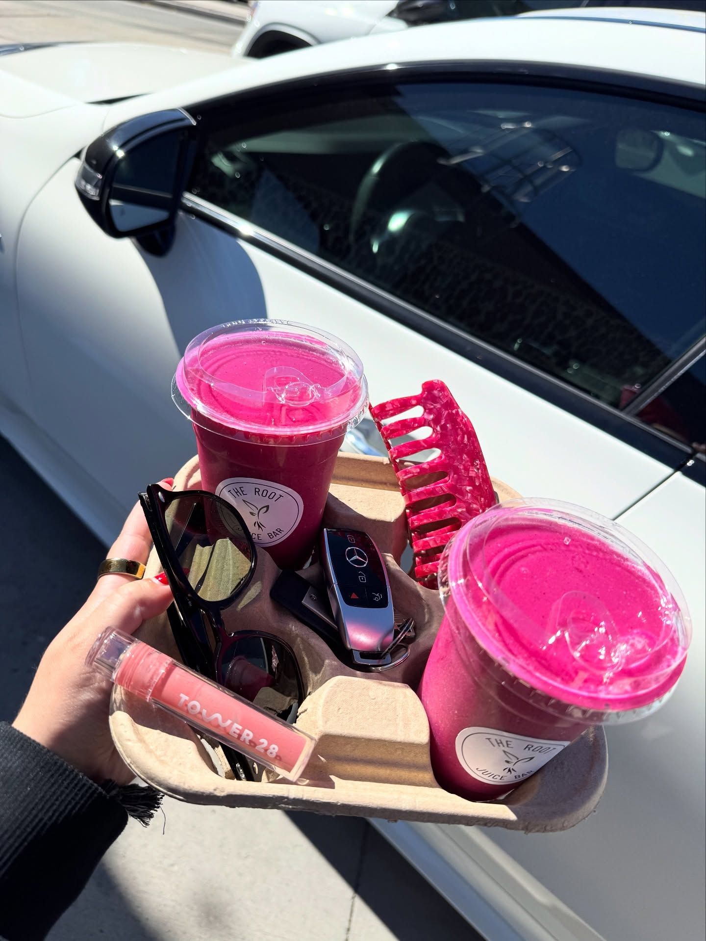 A hand holds a drink carrier with two bright pink smoothies, sunglasses, a car key, lip gloss, and a hair clip by a car.
