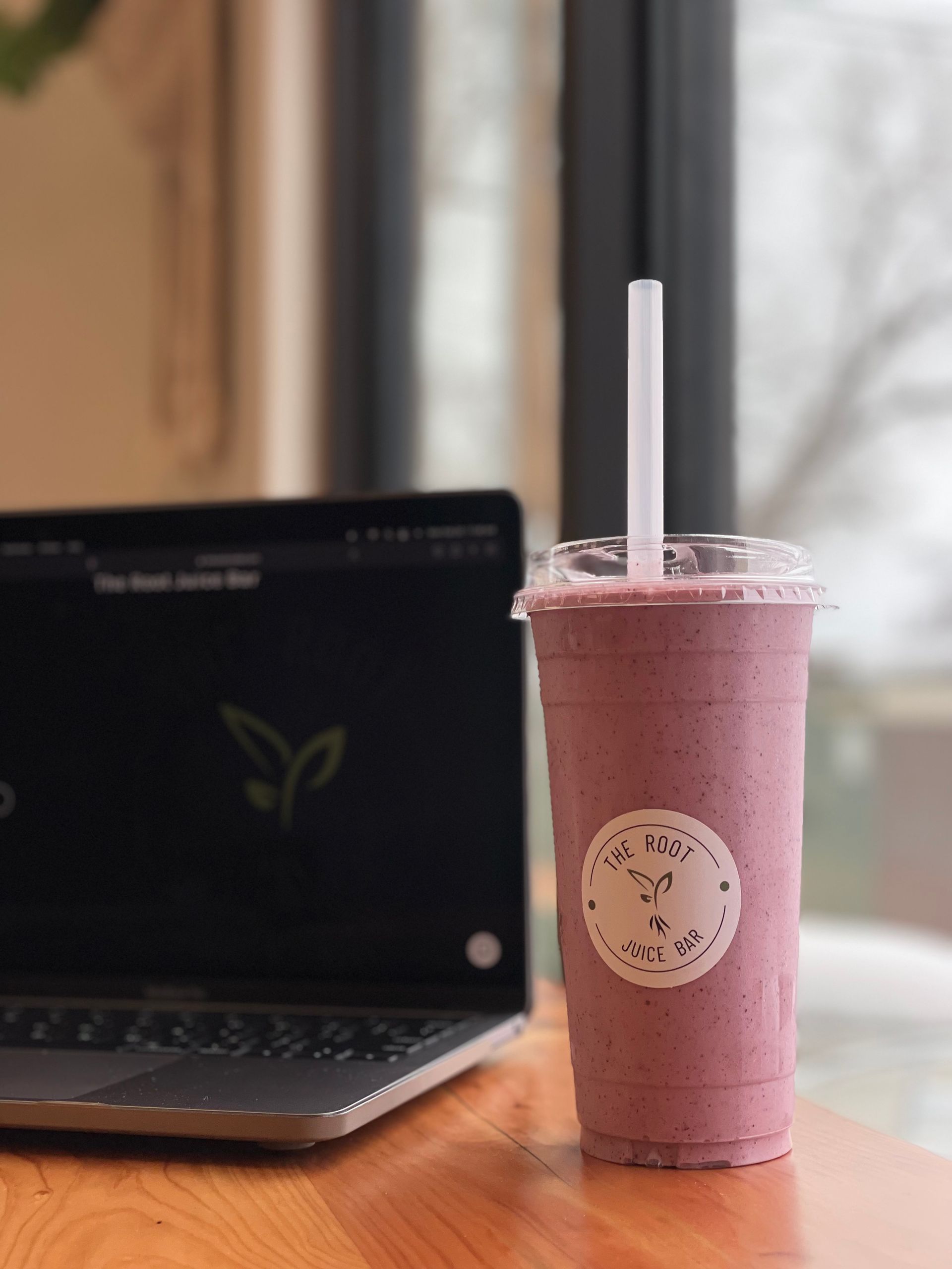 A hand holds an acai bowl topped with granola and berries in front of The Root Juice Bar storefront.