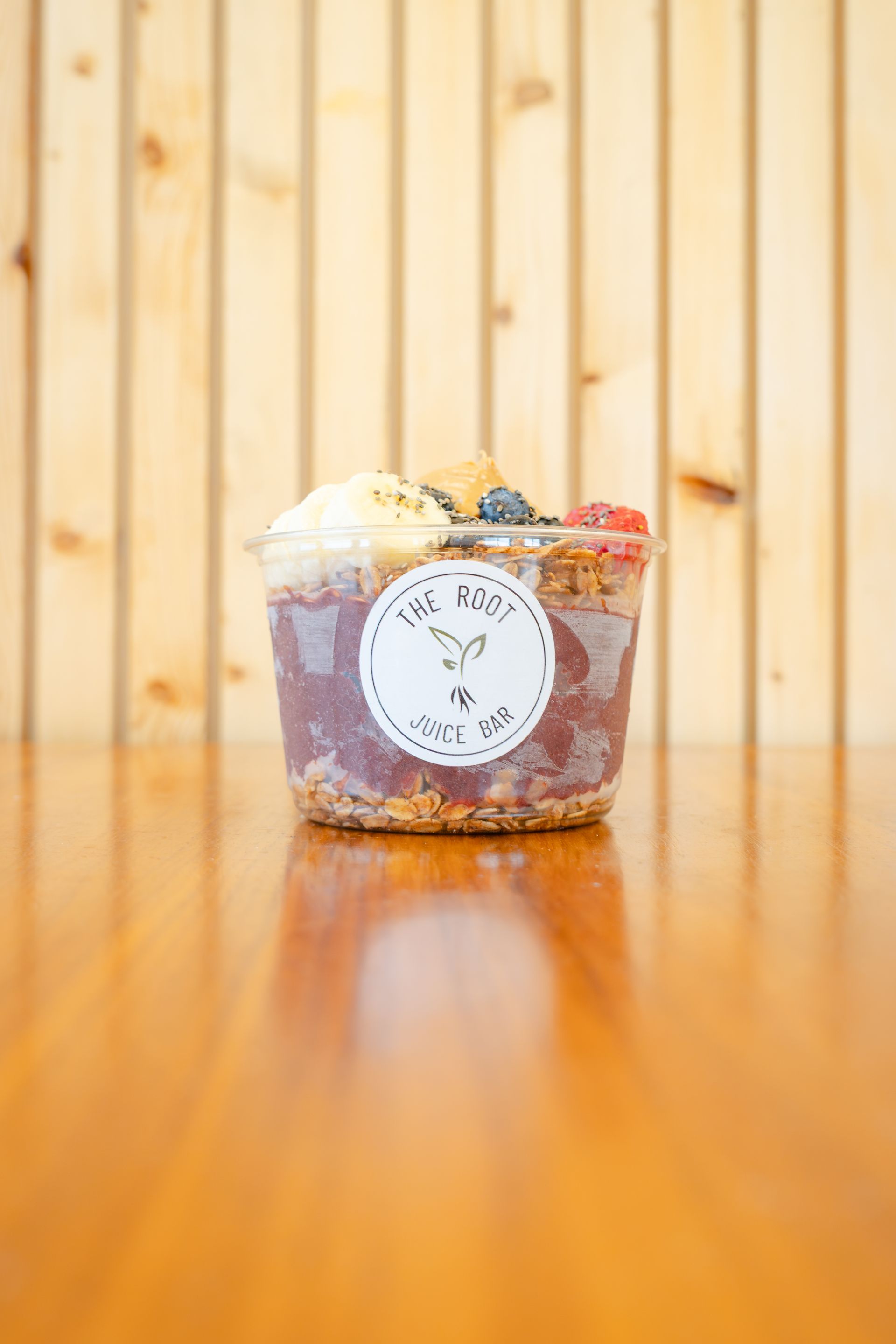 An acai bowl in a clear plastic cup with fruit toppings sits on a wooden table in front of a vertical wood panel wall.