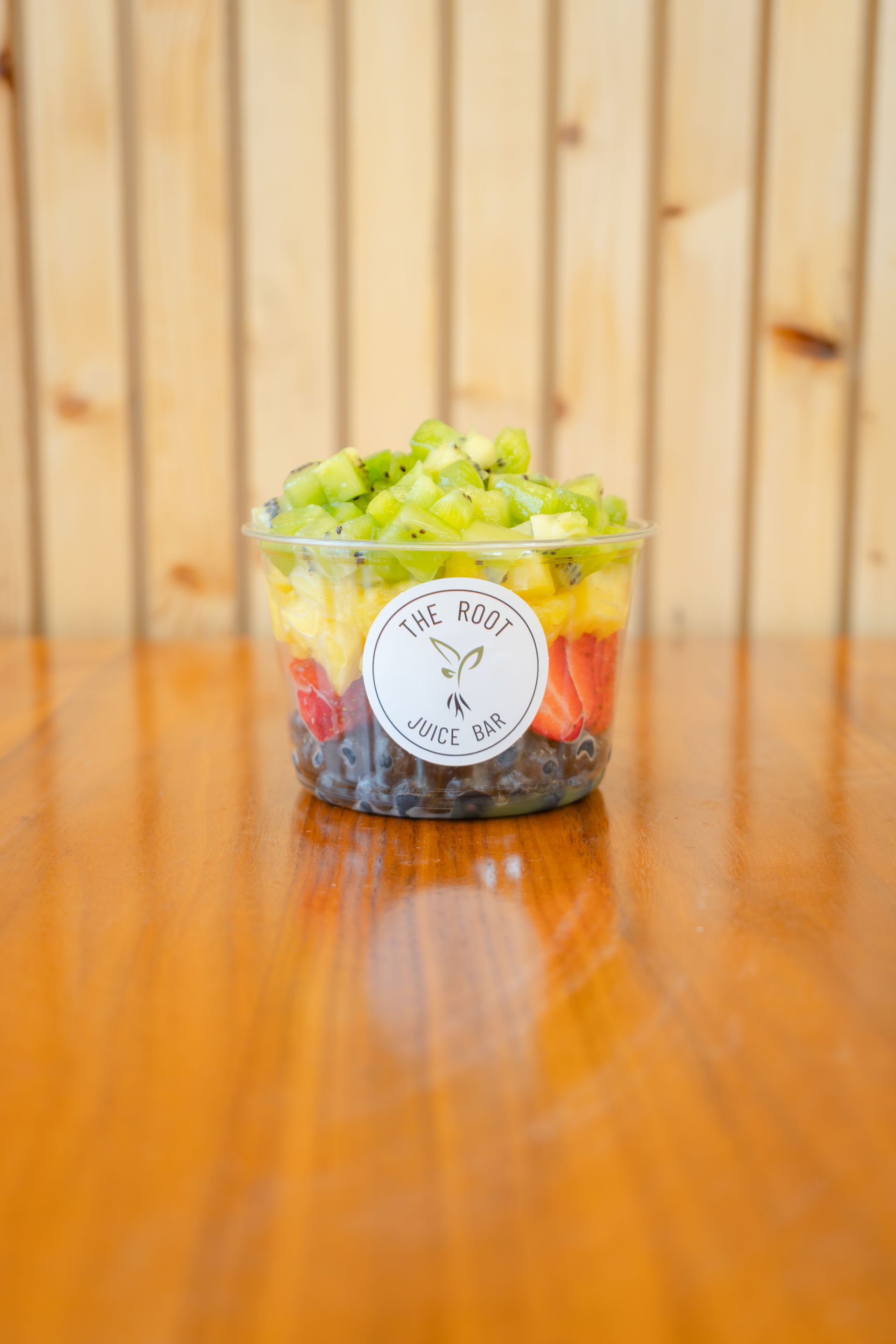 A clear plastic cup of layered fruit, topped with kiwi, sitting on a wooden surface in front of wood-paneled walls.