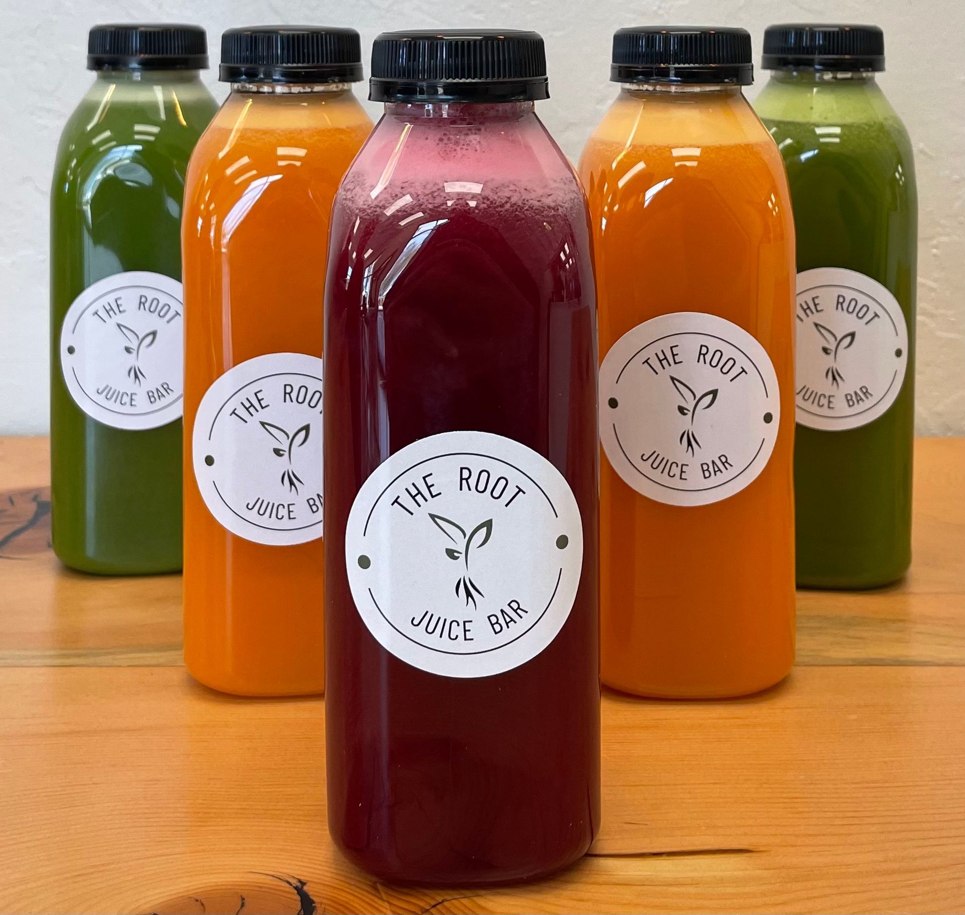 Five bottles of juice from The Root Juice Bar in green, orange, and purple, arranged on a wooden table.