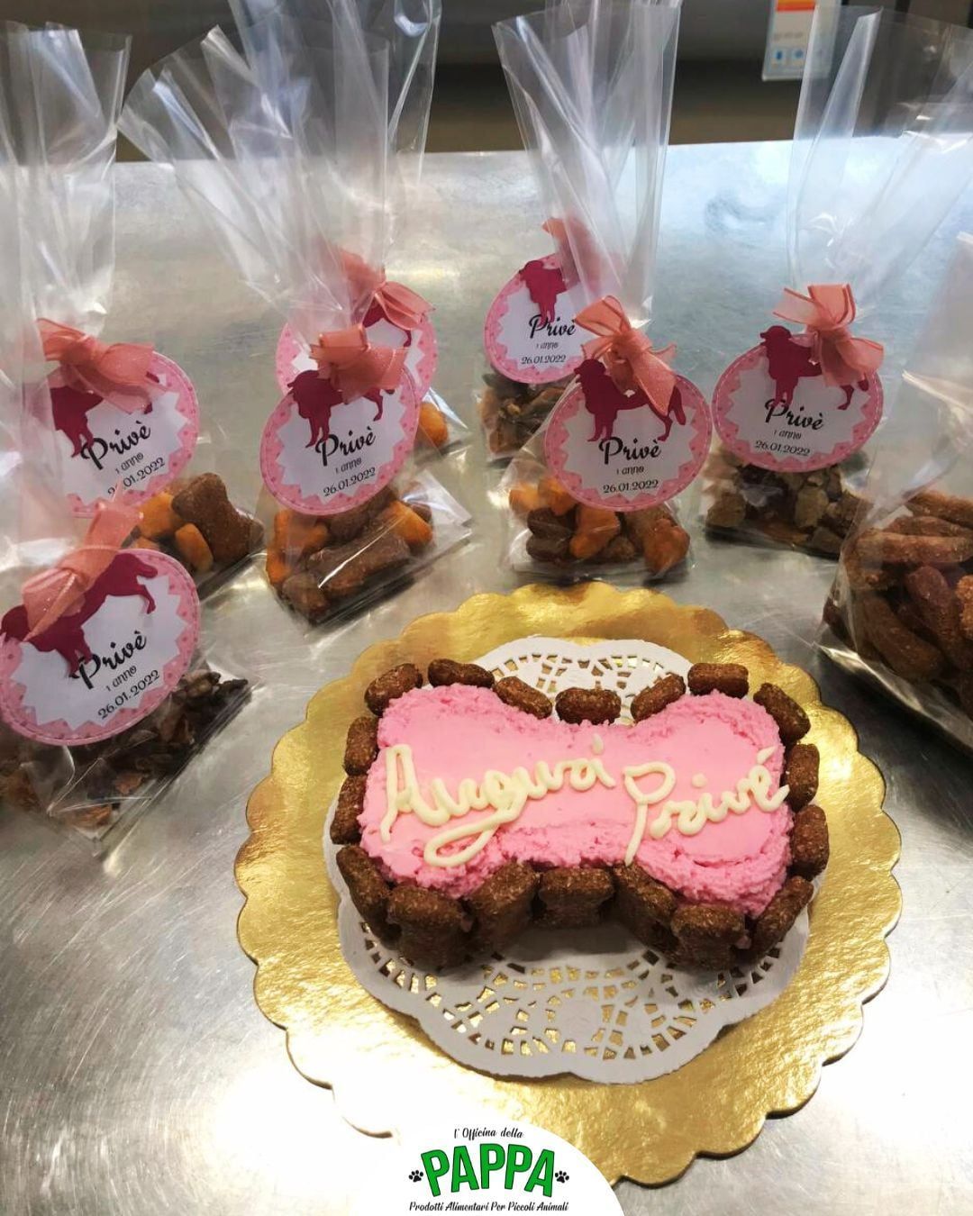 Dolcetti per il compleanno del cane: una torta rosa a forma di fiocco e sacchetti per dolcetti con etichette rosa e dettagli dorati.