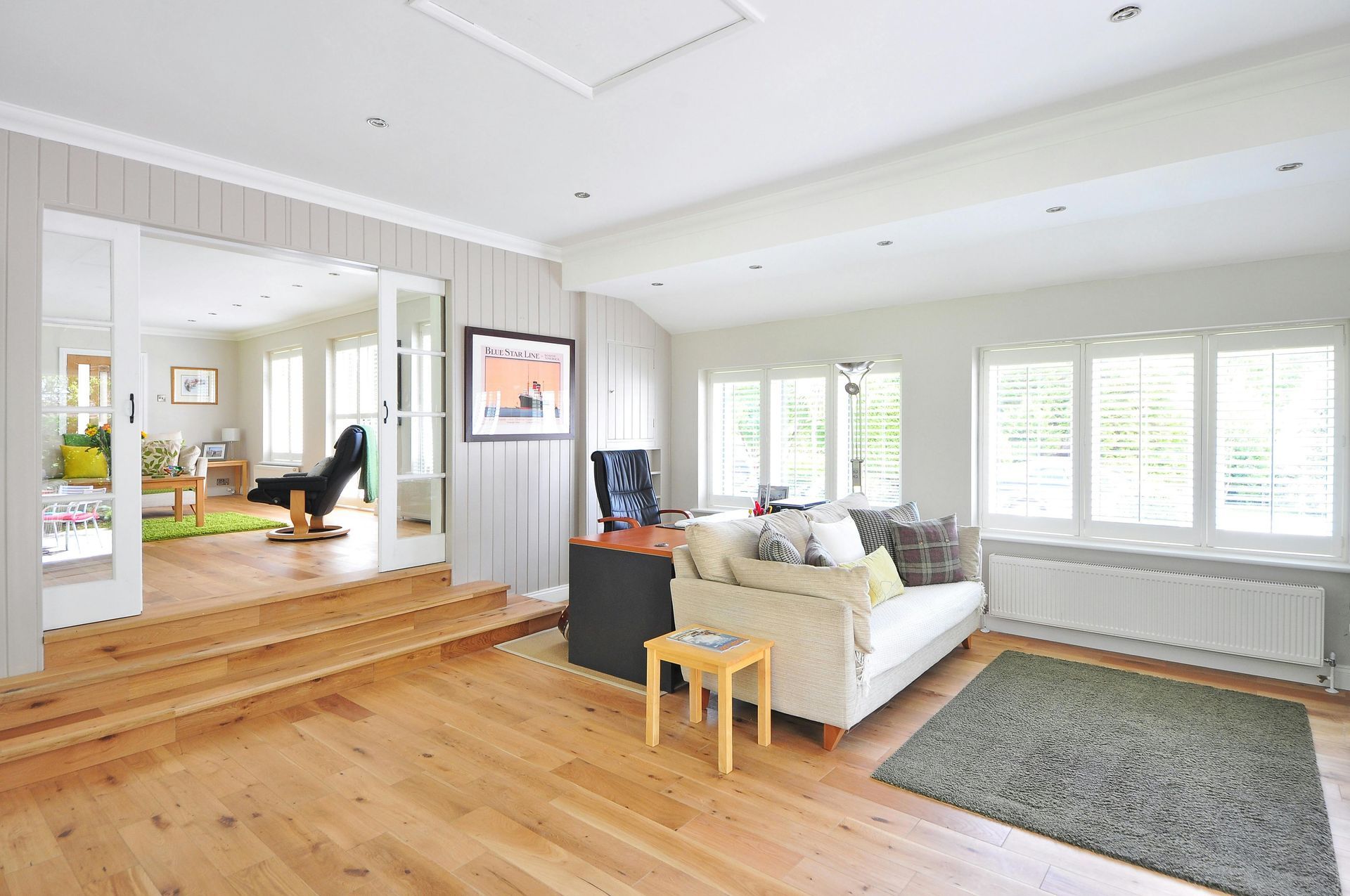 Bright living room with white sofa, wood floor, large windows, and a raised dining area in the background