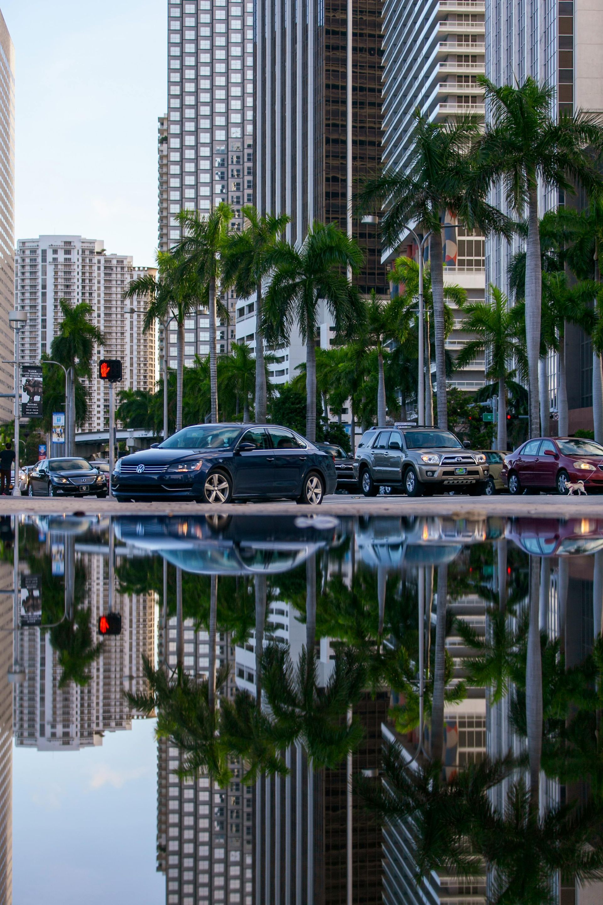 A city street with cars driving down it and buildings reflected in the water.
