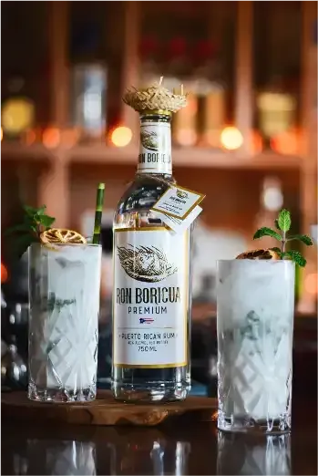 A bottle of Ron Boricua is sitting on a wooden table next to two glasses of mojito drink.