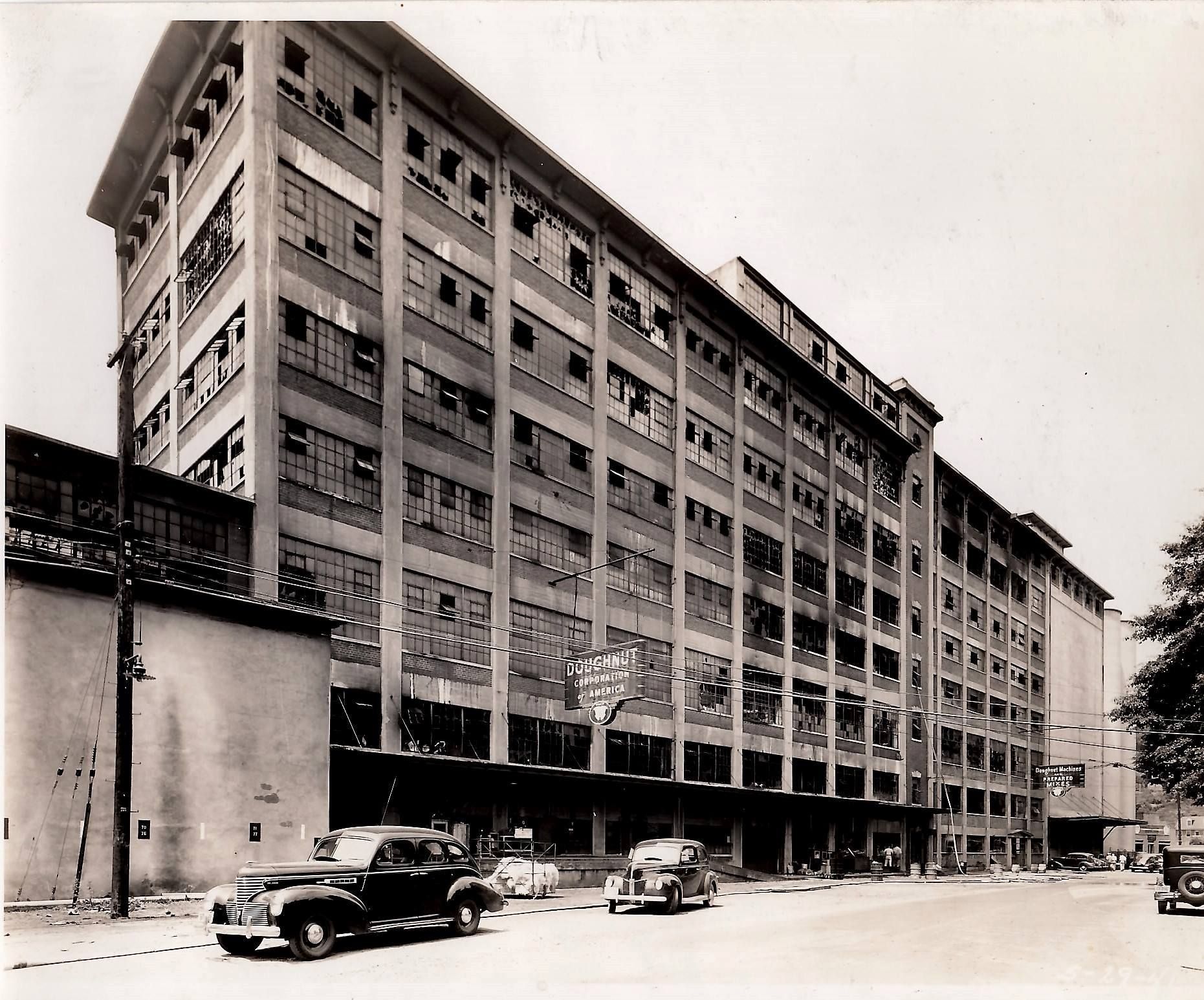 Black-and-white street view of a large multi-story industrial building with parked cars along the curb.