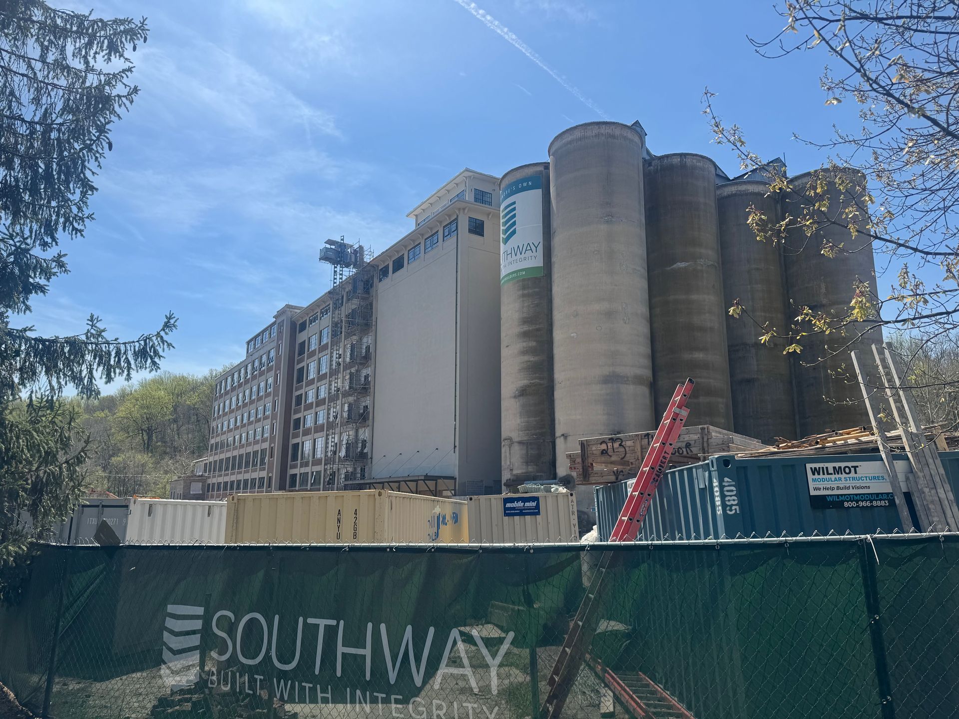 Construction site with Southway fencing, multi-story building, and tall silos under a blue sky