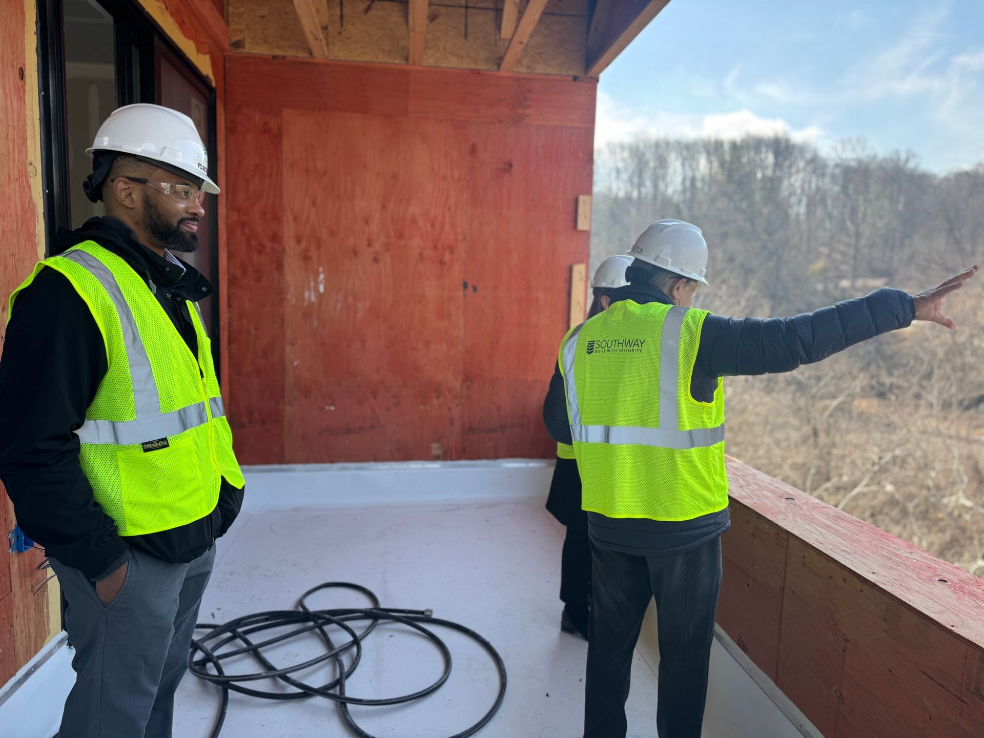 Two workers in hard hats and safety vests stand on a wooden construction platform, one pointing outward.