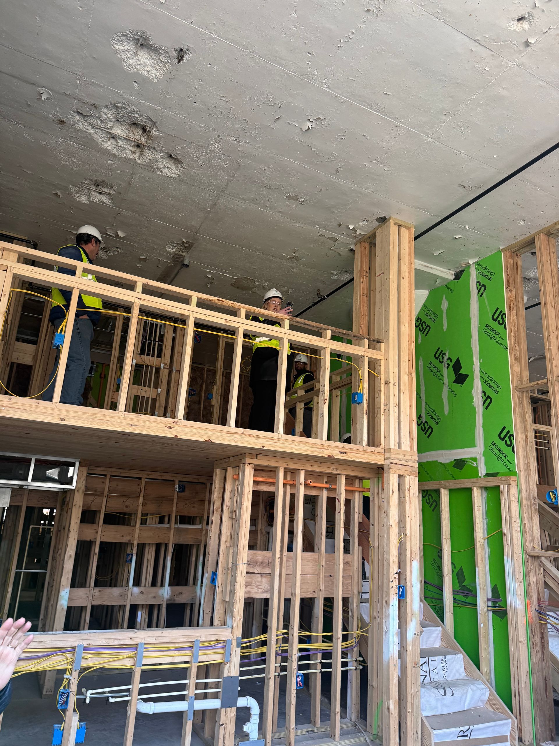 Wood-framed interior under construction with workers on a scaffold and green wall insulation panels.