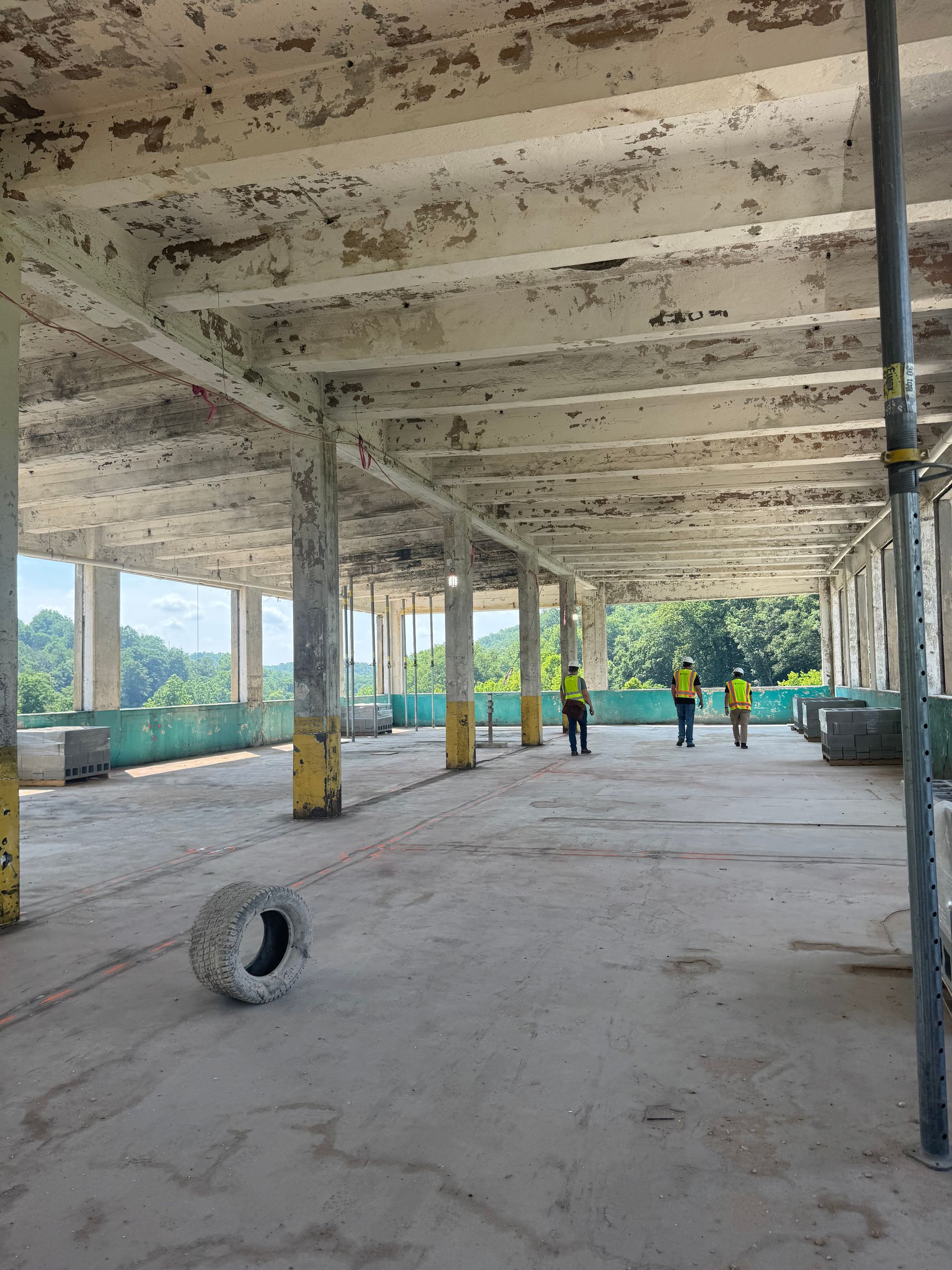 Abandoned concrete building interior with workers in yellow vests near open windows and columns.
