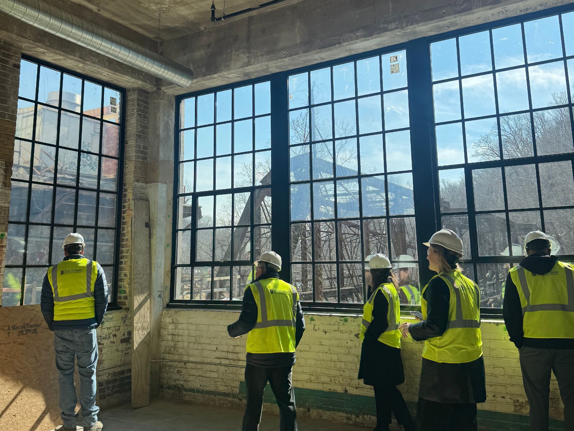 Five workers in yellow safety vests stand in a large industrial building with tall windows.