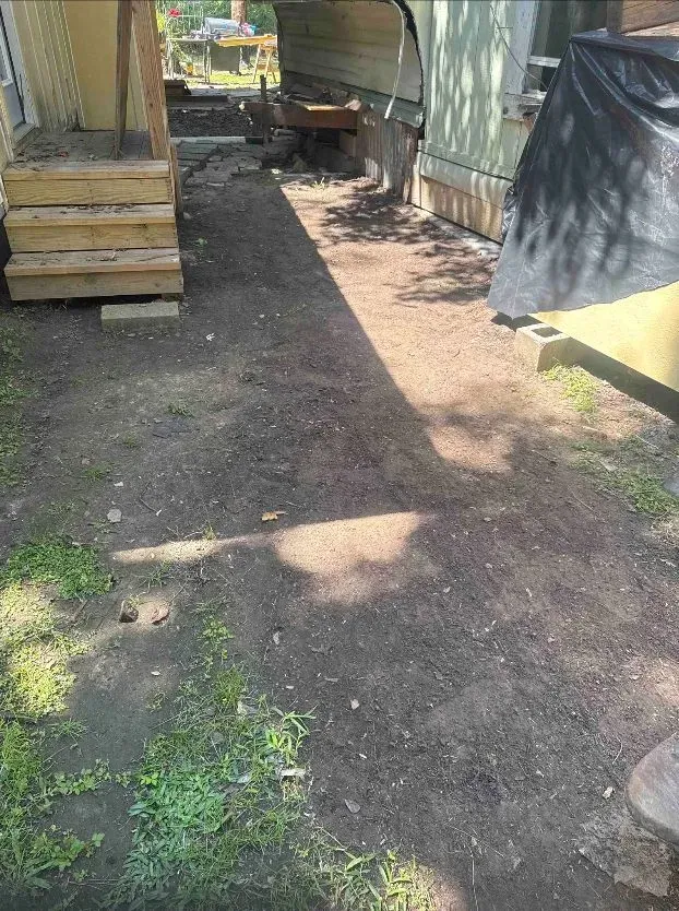 Dirt path between two buildings, wooden steps on the left. Green grass and shadow on the ground.