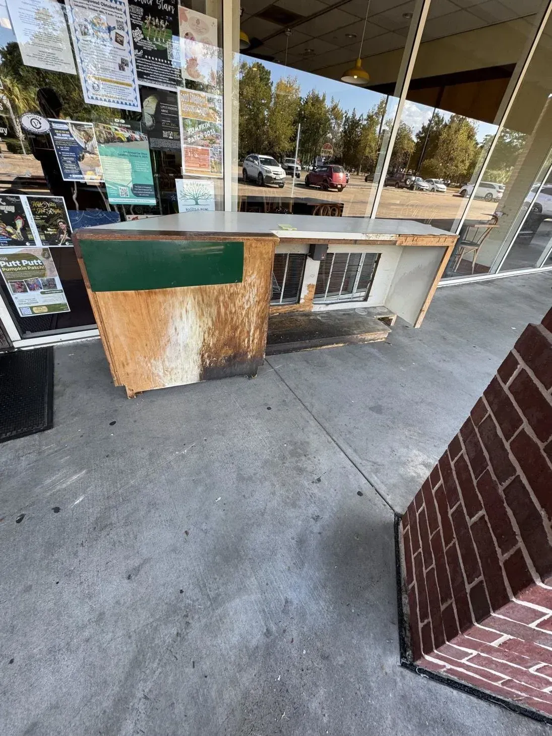 Weathered, damaged counter outside a storefront, with glass windows reflecting vehicles and trees.