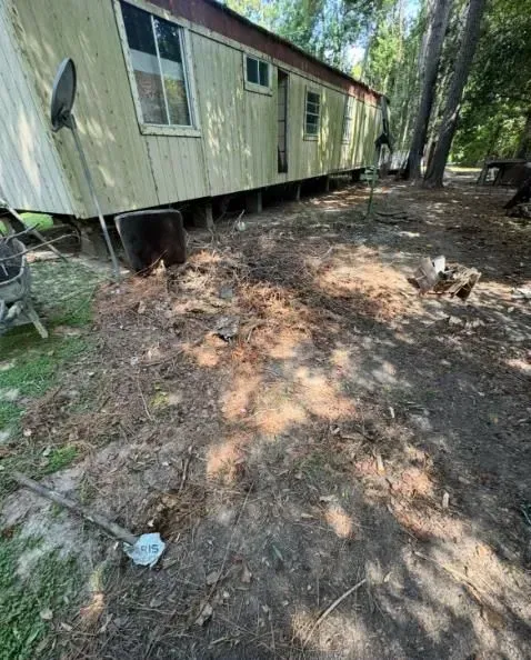 Mobile home exterior with debris on ground, shaded by trees, with satellite dish and shovel.