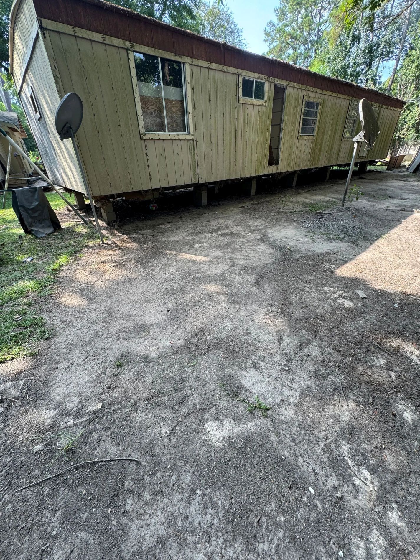 A worn mobile home with a satellite dish, set on wooden blocks on a dirt lot.
