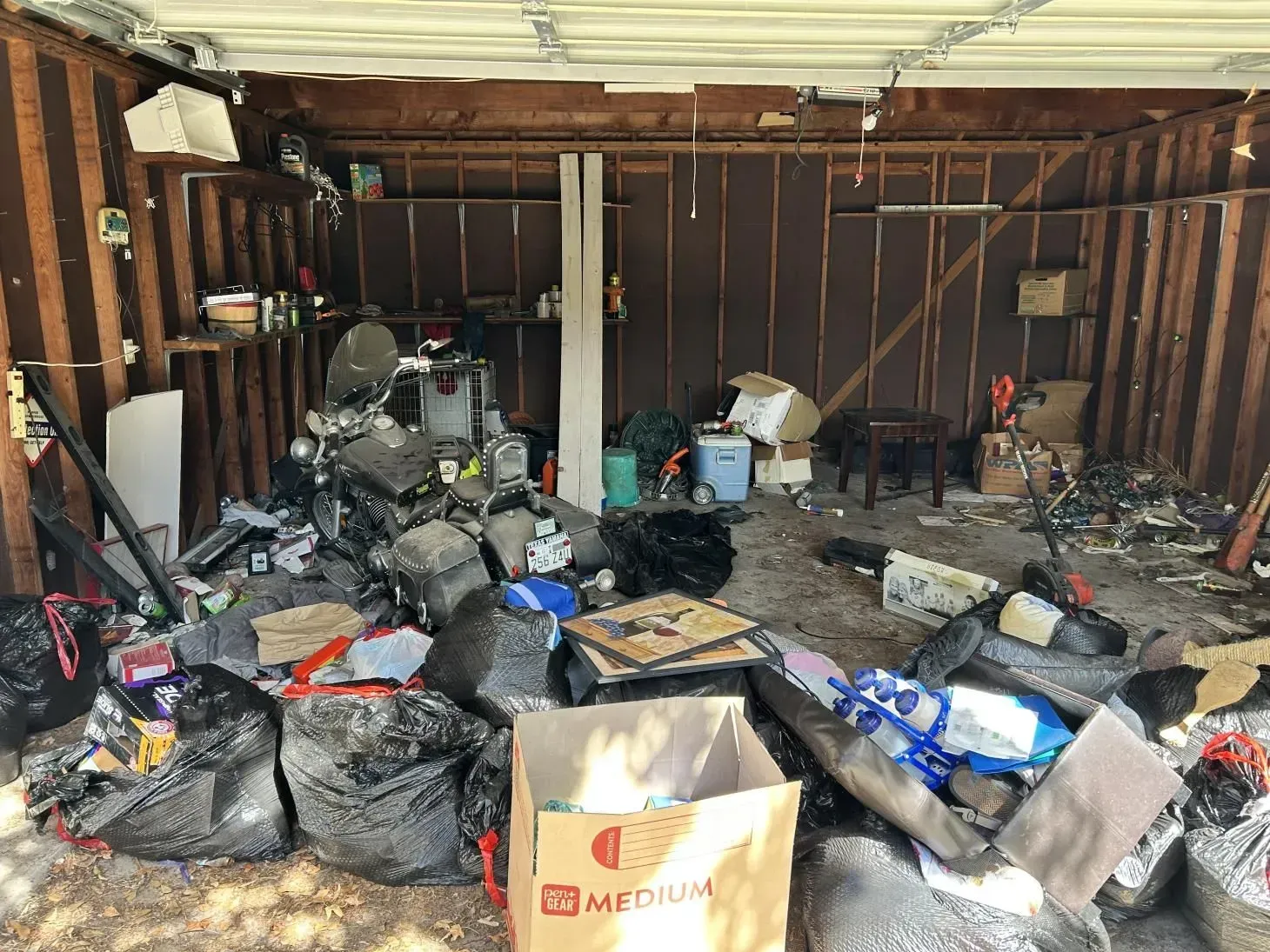 Cluttered garage with trash bags, boxes, and debris scattered across the floor and shelves. Wooden structure.