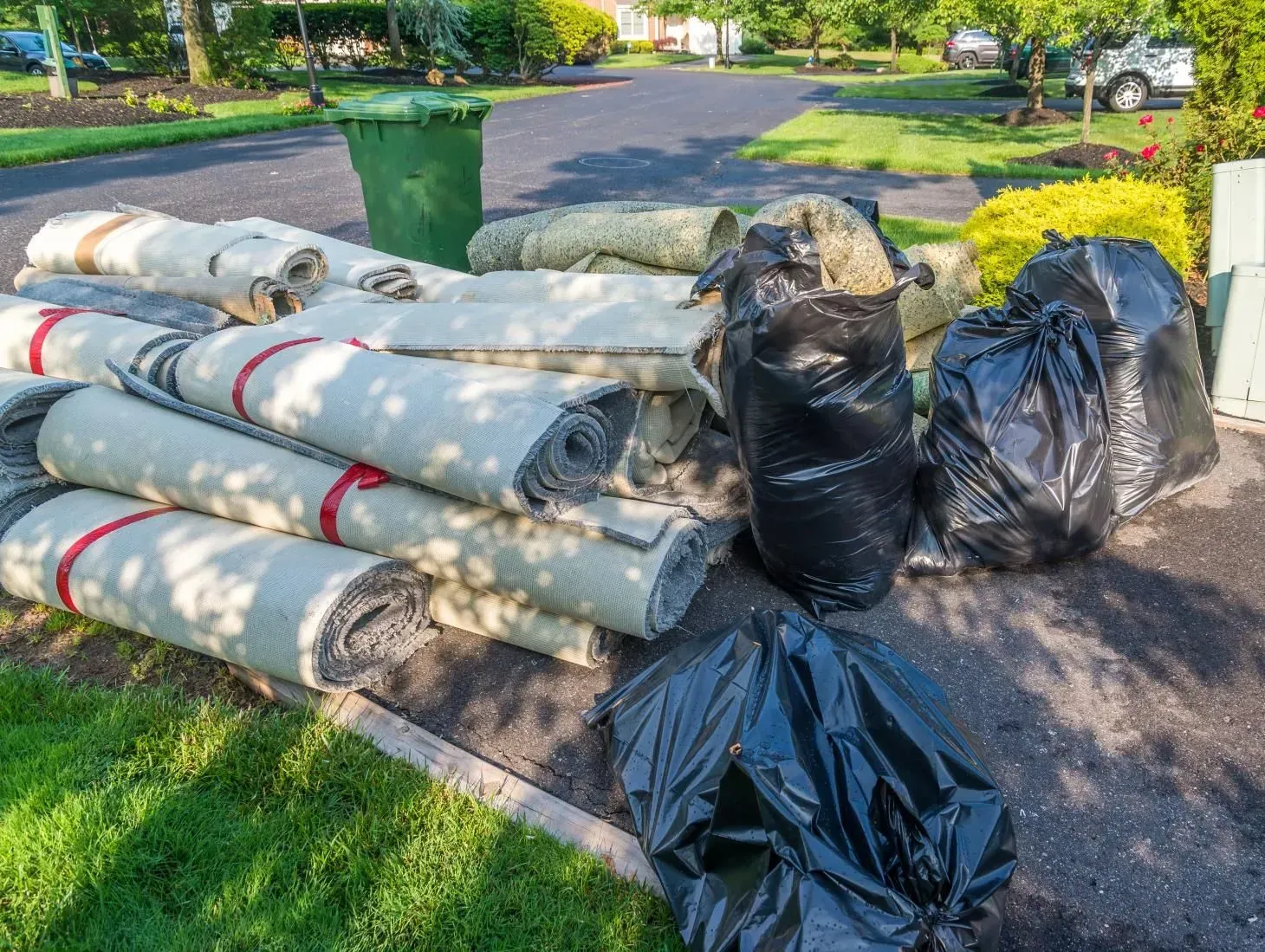 Rolls of old carpet and garbage bags piled on a driveway next to a green trash can, outside.