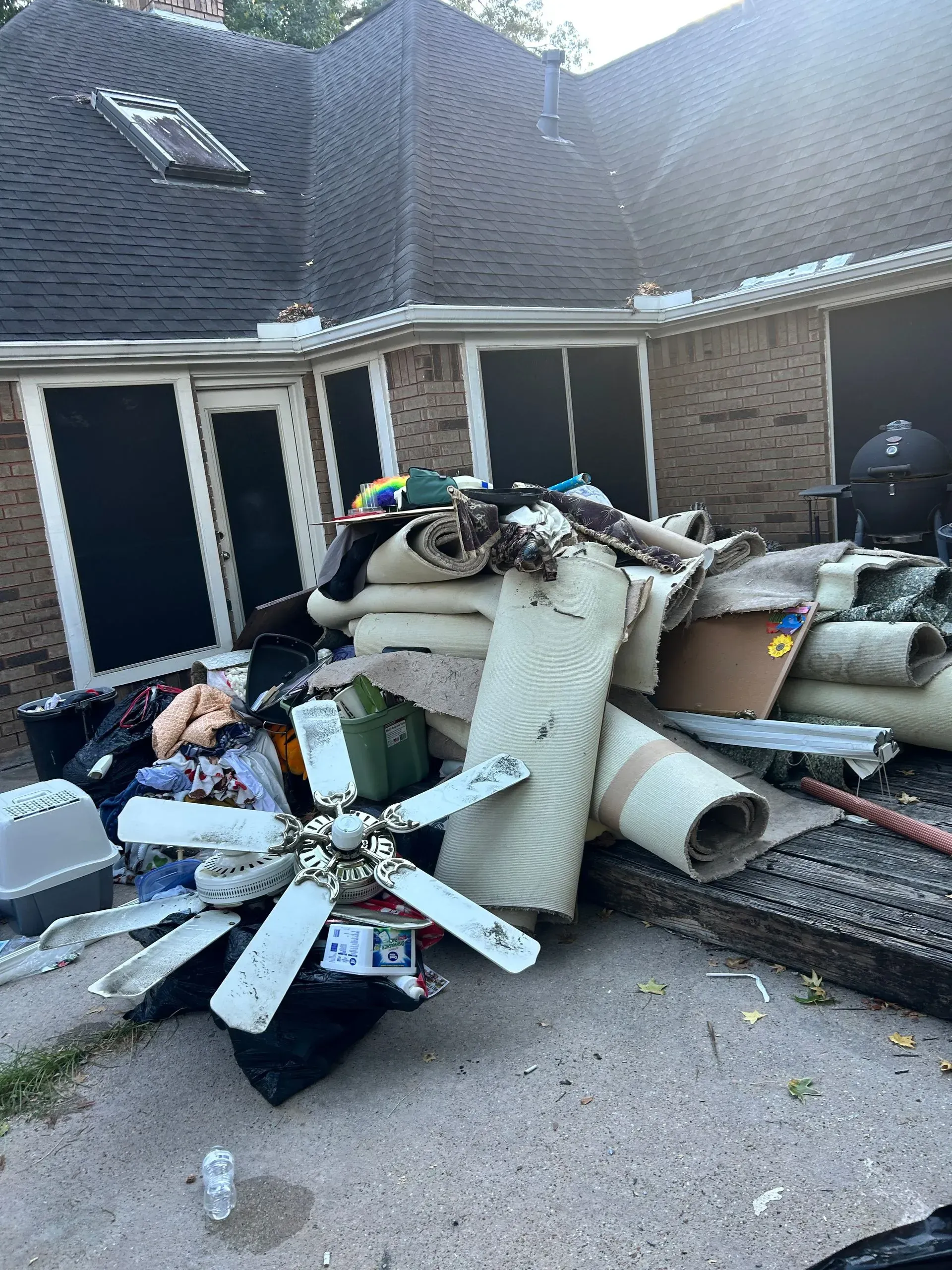 Pile of debris including carpet, fan, and trash bags next to a house with dark roof and windows.