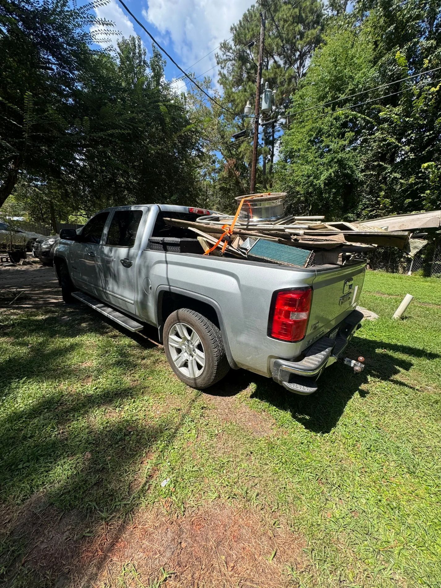 Silver pickup truck bed filled with lumber, parked on a grassy area. Trees and sky in the background.