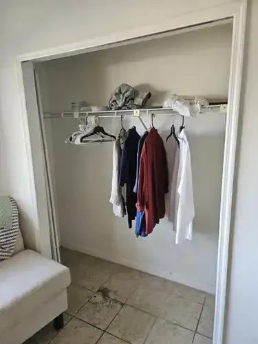 A small, white-framed closet with clothes hanging on a rod. Sneakers and a cap on shelf above.