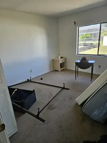 Empty bedroom with bed frame, nightstand, table, and window overlooking a yard.