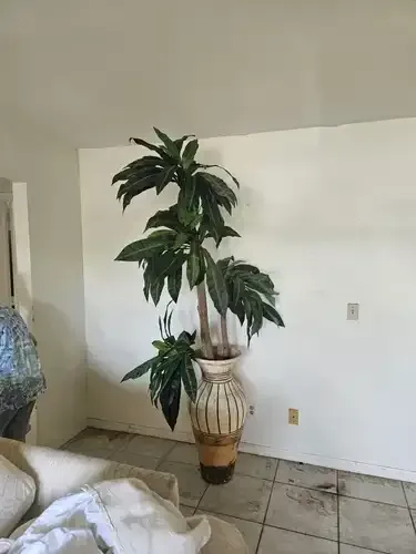 Tall artificial plant in a decorative vase against a white wall in a room.