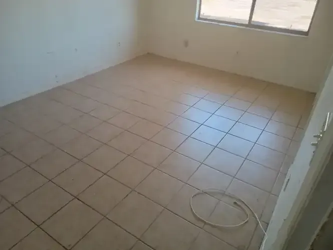 Empty room with beige tiled floor, white walls, and a window. A white cord is visible on the floor.