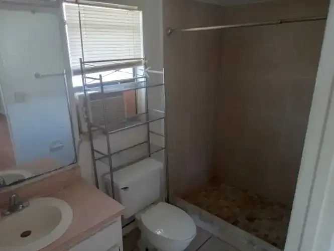 Bathroom with toilet, sink, shower, and window. Pink counter, metal shelving, and beige walls.