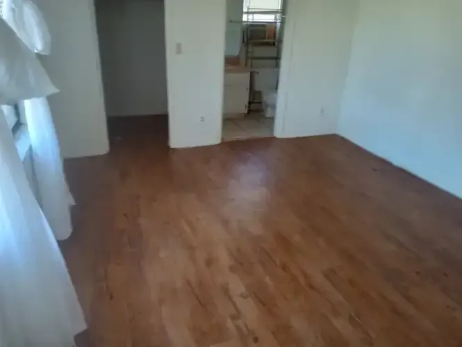 A room with wood floors and white walls. Another room and a doorway are visible.