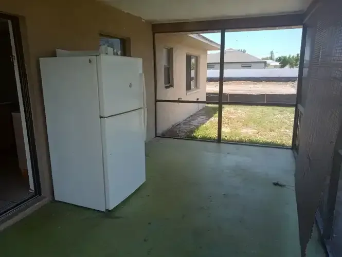 White refrigerator on a green screened-in porch, with view of a yard and houses.
