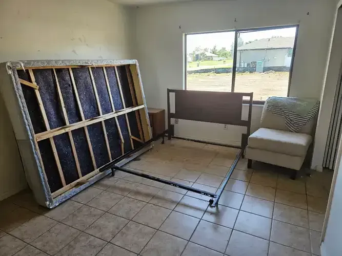 A mostly empty bedroom with an unassembled bed frame, mattress, chair, and a nightstand.