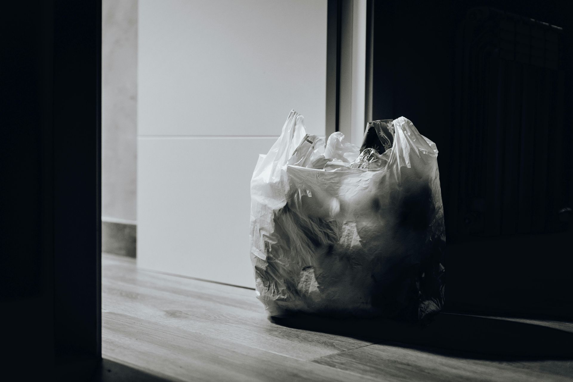 A plastic trash bag filled with garbage sits on a wooden floor next to a closed door.