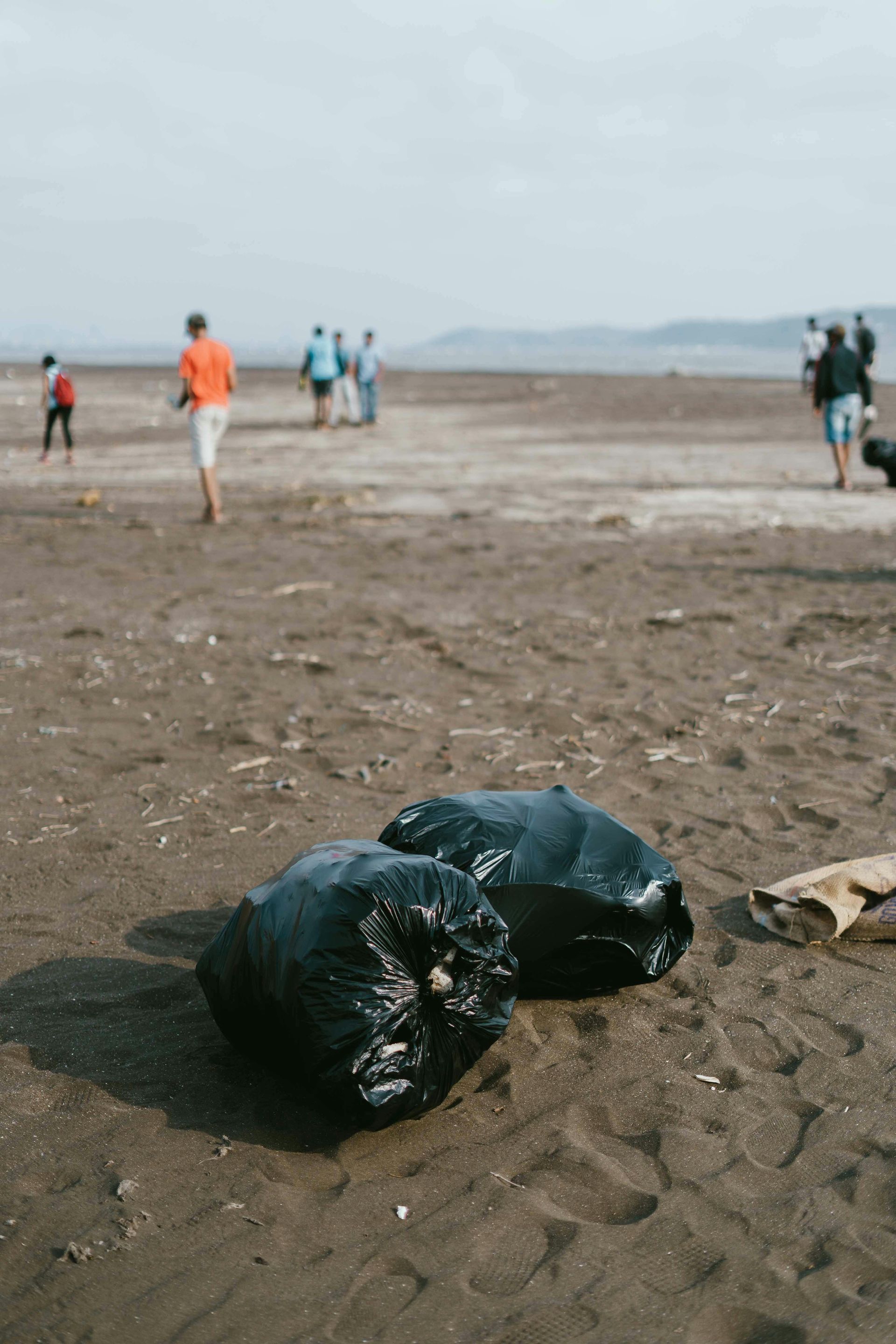Two black trash bags on a sandy beach, people walking in the background.