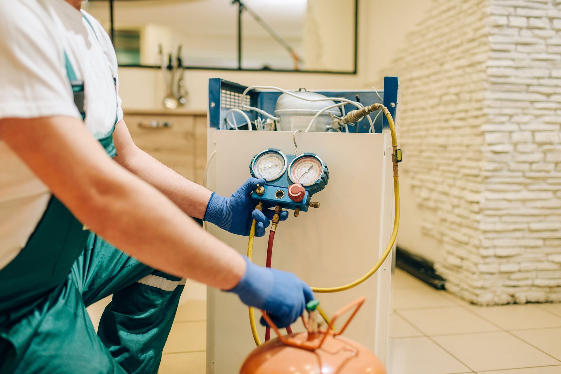 HVAC technician in green coveralls refilling refrigerant with gauges and orange tank in room.