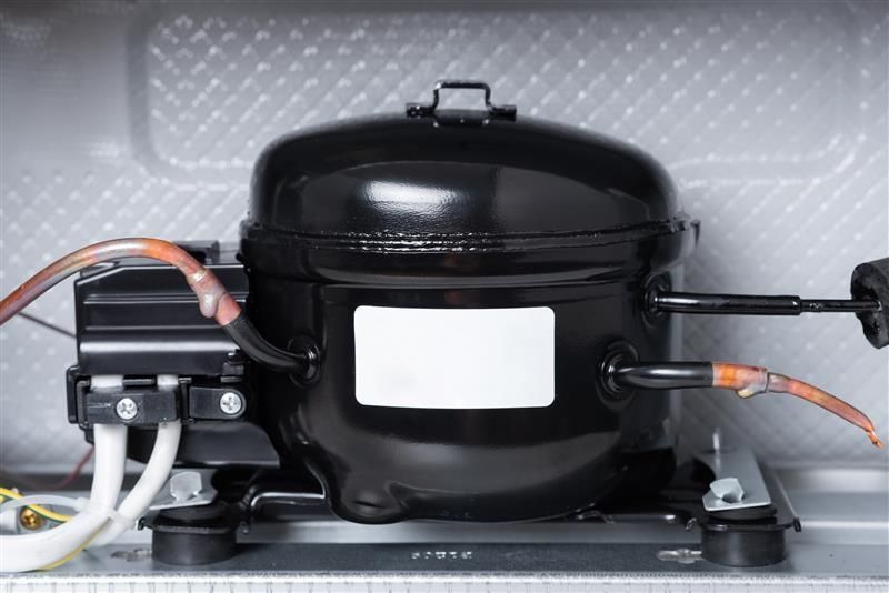 Black refrigerator compressor with copper tubing and electrical connections.