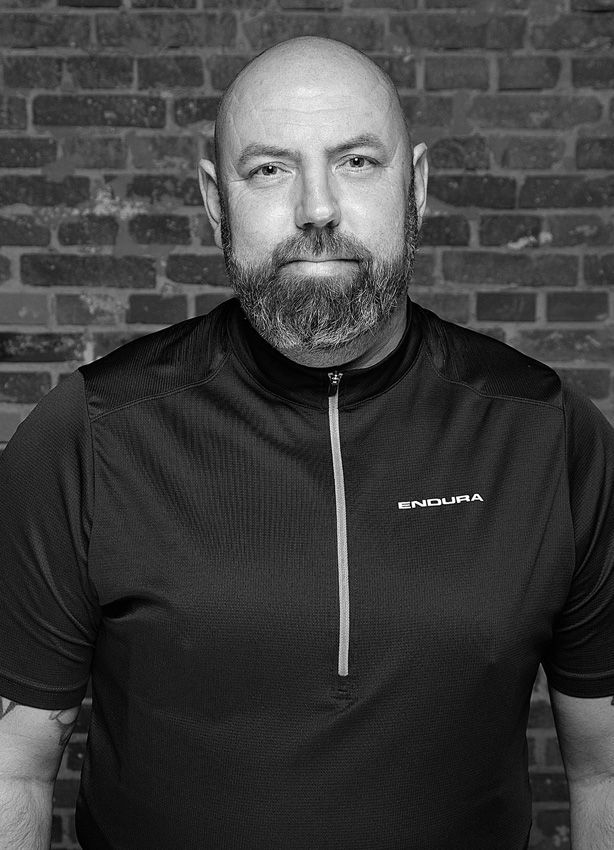 En skaldet mand med skæg iført sort skjorte står foran en murstensvæg.