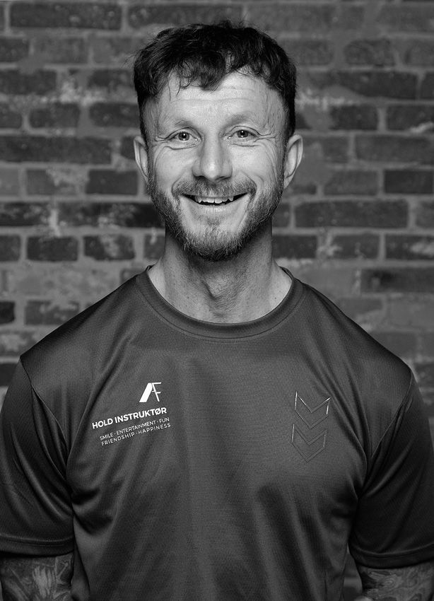 En mand med skæg smiler foran en murstensvæg på et sort-hvidt foto.