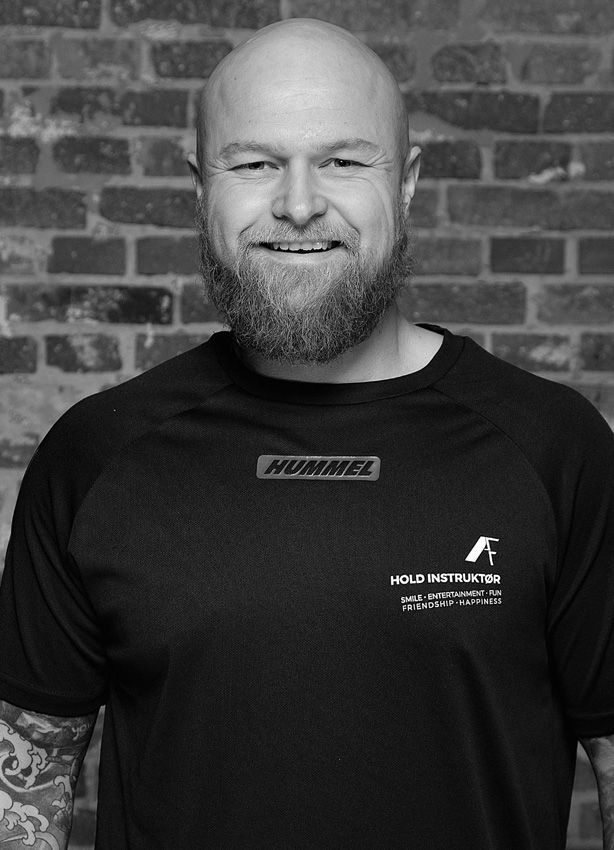 En mand med skæg er iført en sort skjorte og smiler foran en murstensvæg.