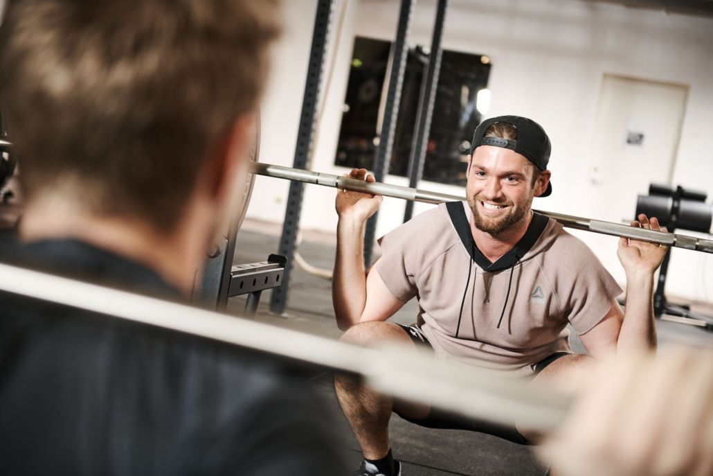 En mand sidder på hug med en vægtstang i et fitnesscenter, mens en anden mand ser på.