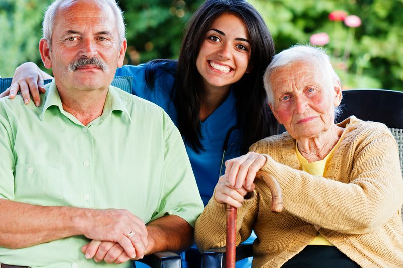 A caregiver smiling behind an older man and woman, one holding a cane, seated outdoors.