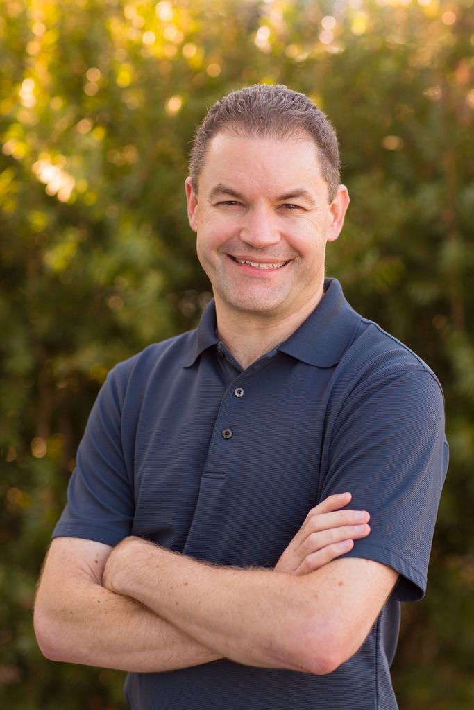 A man in a blue shirt is standing with his arms crossed and smiling.