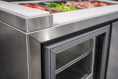 Stainless steel refrigerated food prep counter with a cutting board top and open door showing shelves of food containers.