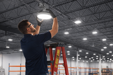 A person in a dark shirt and safety gloves standing on a red ladder while repairing a bright overhead warehouse light.