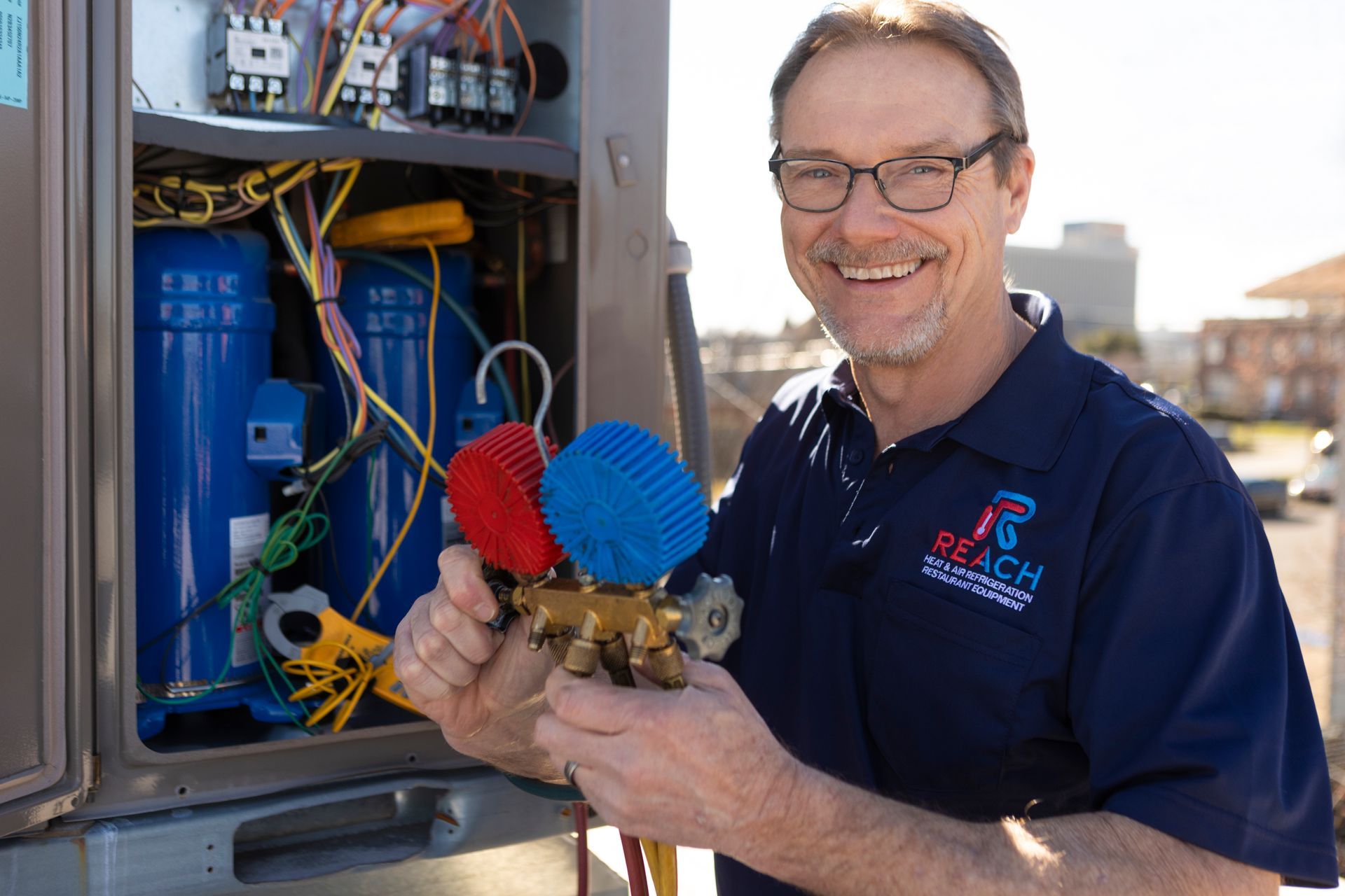 A technician smiles while holding a set of blue and red HVAC manifold gauges in front of an open air conditioning unit.