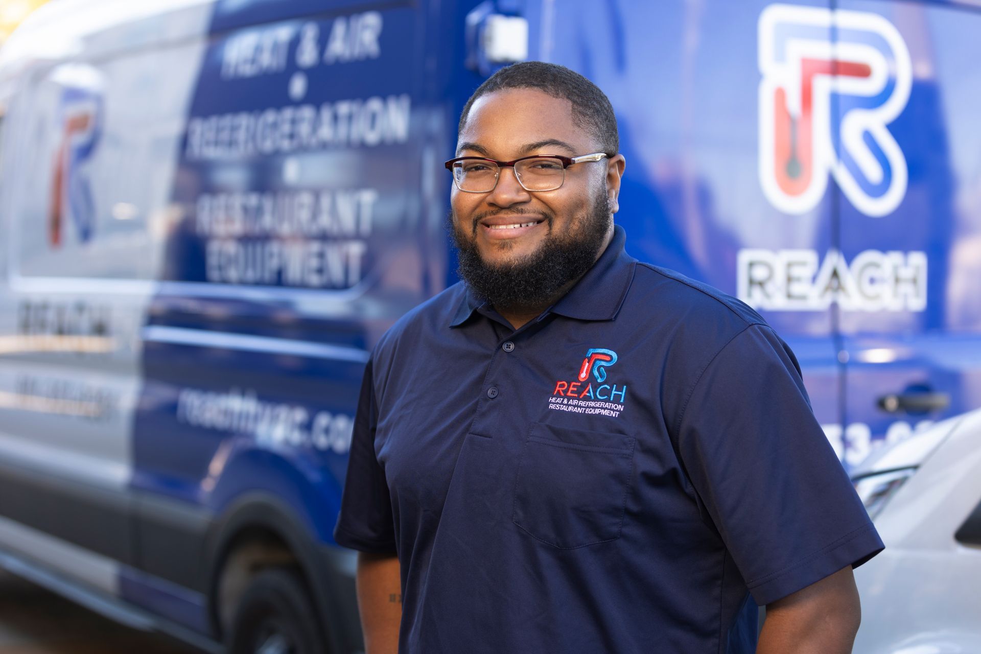 A person in a blue Reach HVAC uniform smiling in front of a company van with branding visible on the vehicle.
